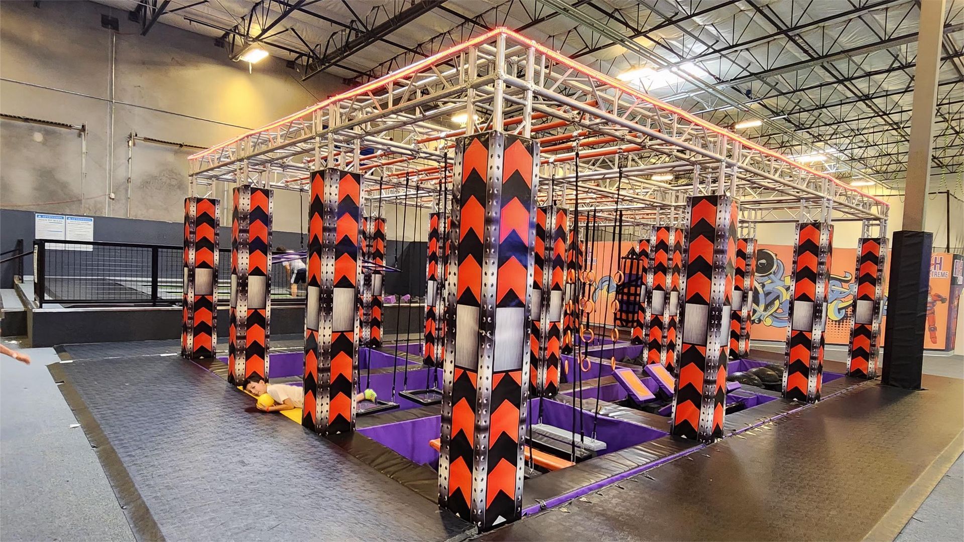 Indoor obstacle course with trampolines, metal scaffolding, and orange/black padding.