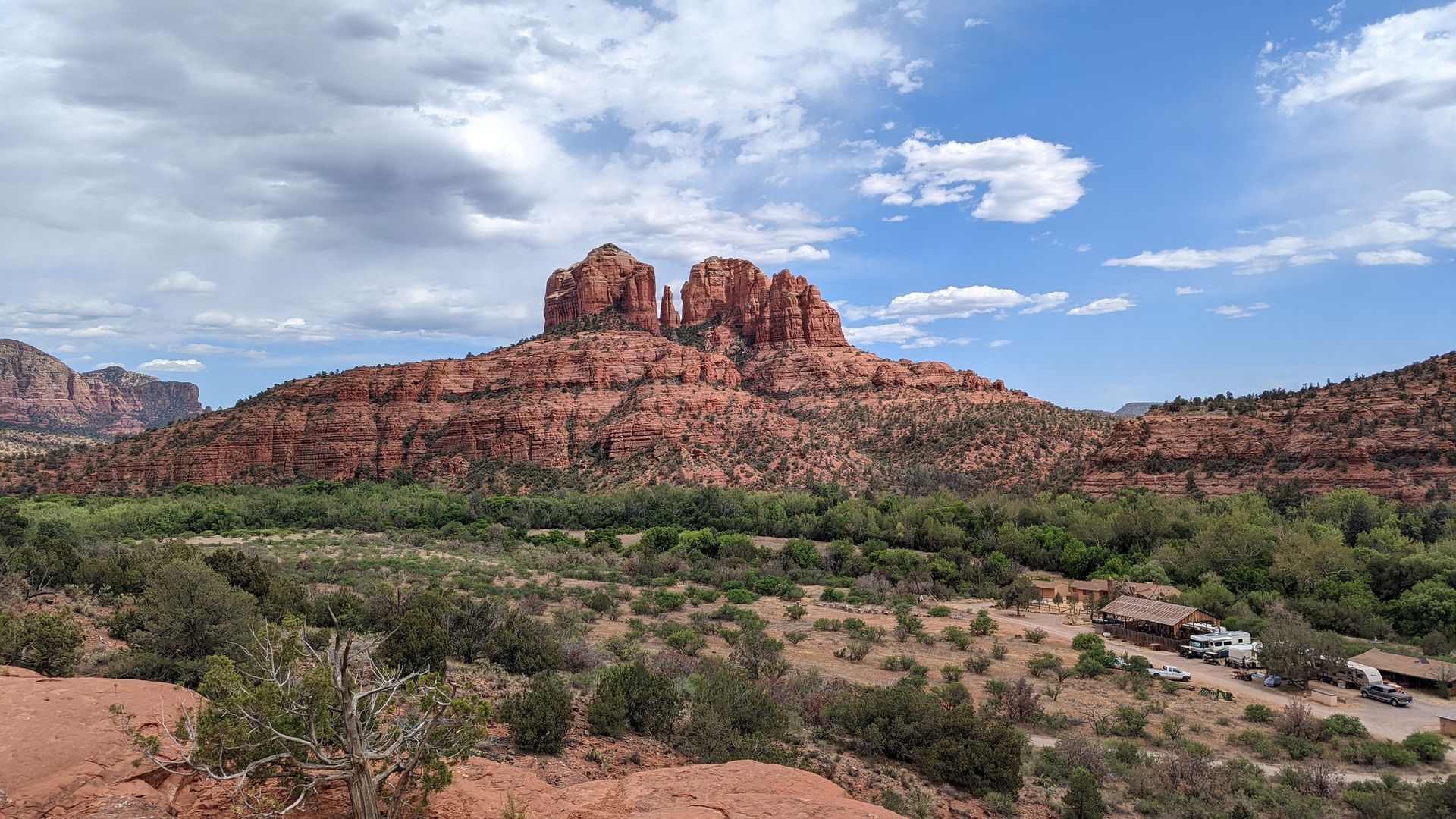 Red rock butte towers over a green valley under a cloudy blue sky in Sedona, Arizona.