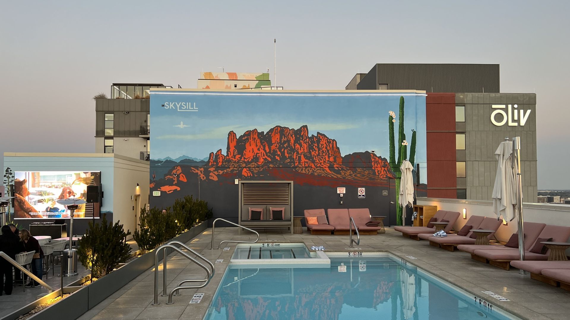 Rooftop pool with desert mural backdrop; people lounge on chairs, watching a screen.