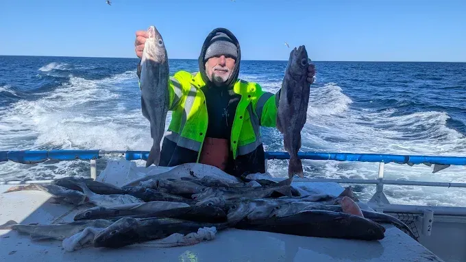 Fisherman on boat holding up fish with a table full of catch; blue water and sky.