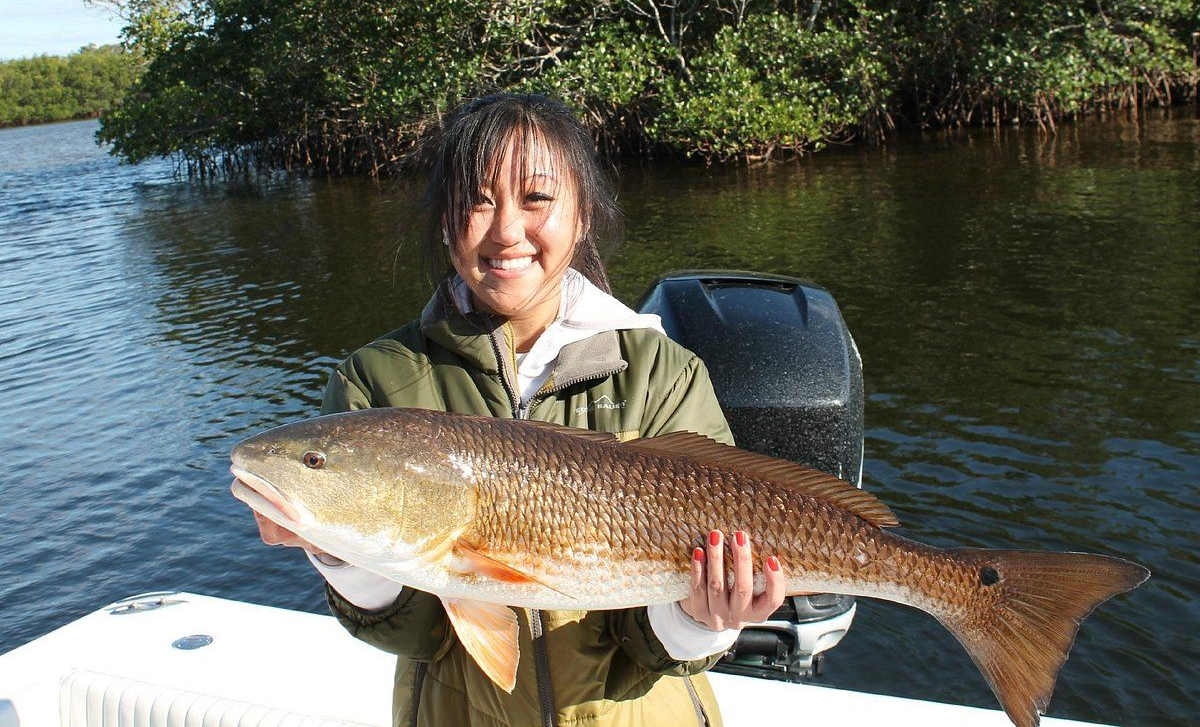 Woman on boat holding a large redfish. Smiling, near mangroves, blue water.