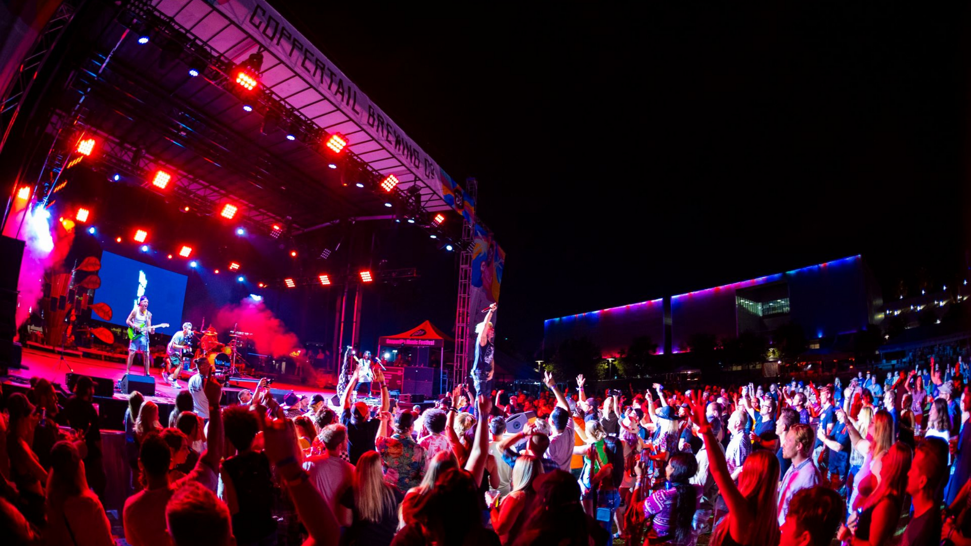 Concert with a band on stage, a large crowd with raised hands, and bright red and blue stage lights at night.