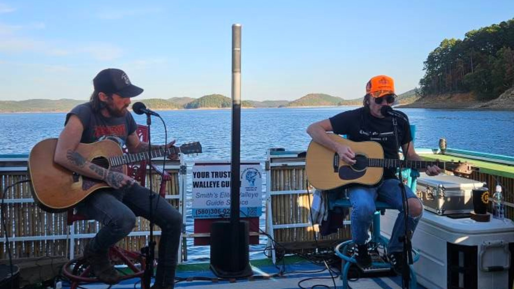 Two guitarists performing live music on a boat, lake in background, under a covered area.