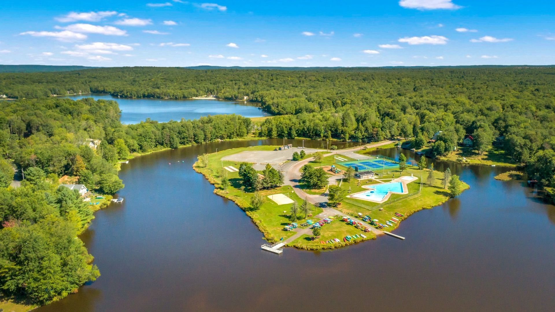 Aerial view of a lake and island with swimming pools, trees, and buildings, under a blue sky.