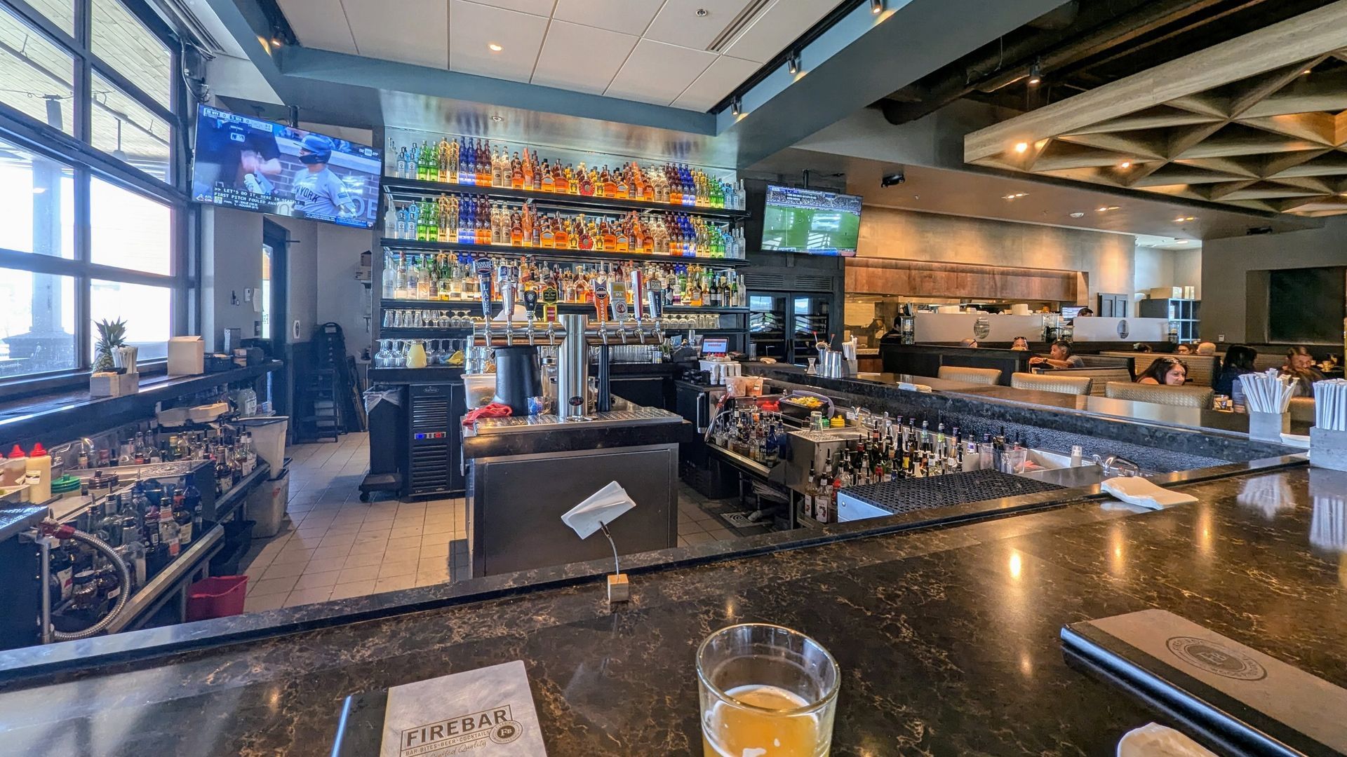 Bar with bottles, taps, and multiple televisions. Empty tables, daytime setting, and a glass of beer on the counter.