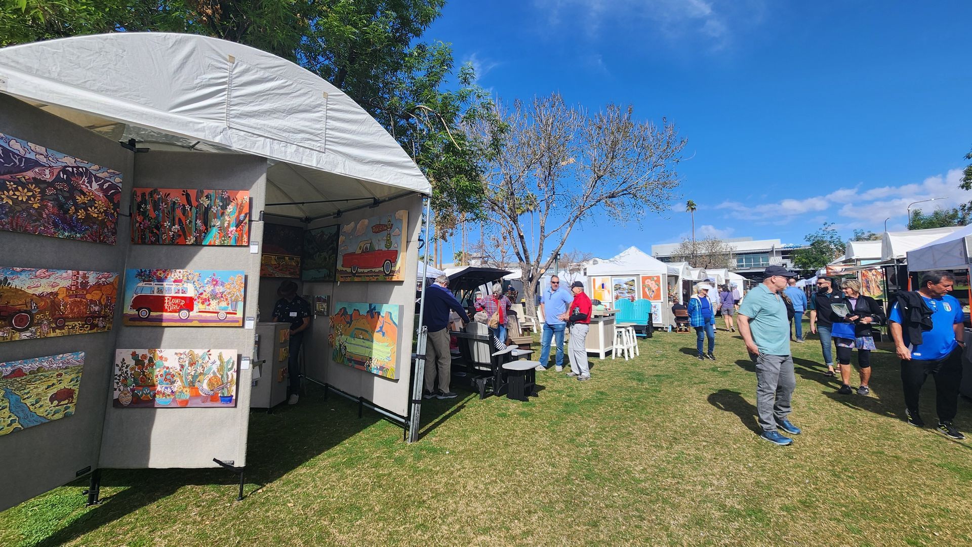 Art fair with white tents, displaying art on grass under a blue sky. People browse and chat.