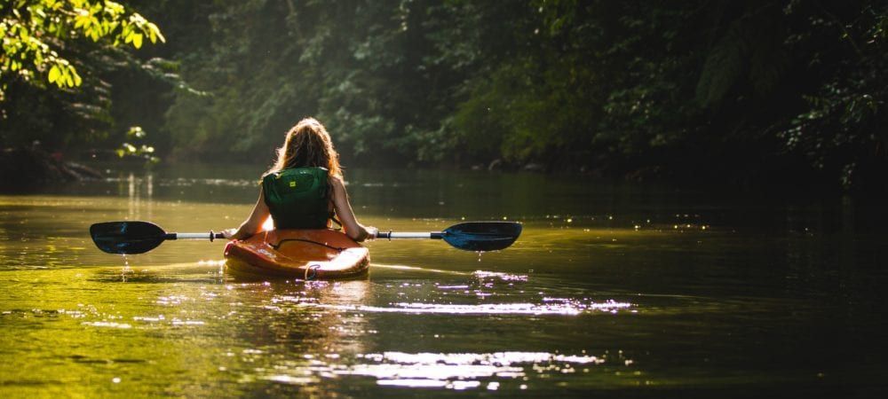 Person paddles a kayak down a sunlit river, surrounded by trees.