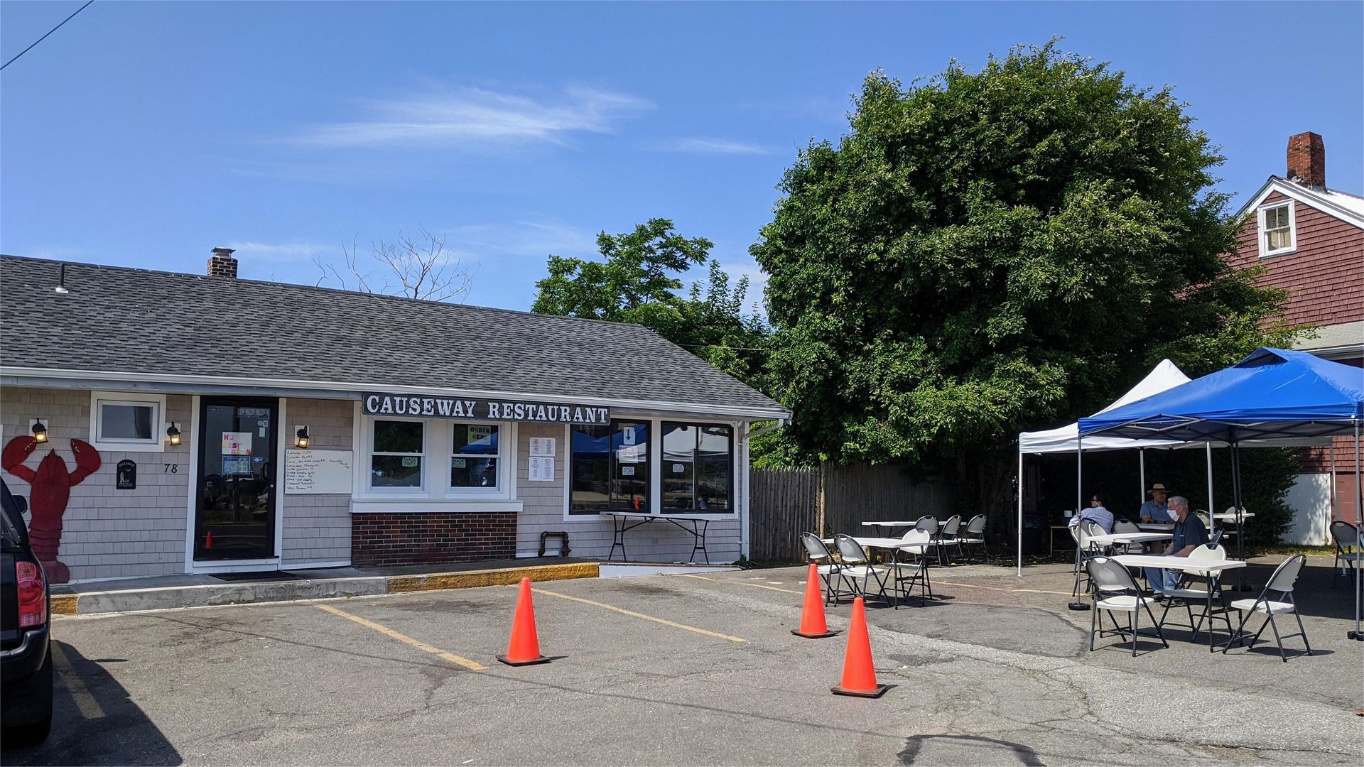 Restaurant exterior with outdoor seating under tents and orange traffic cones in parking area.