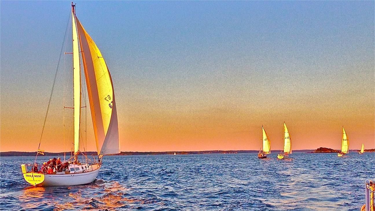 Sailboat with yellow sail on blue water at sunset, followed by other sailboats.