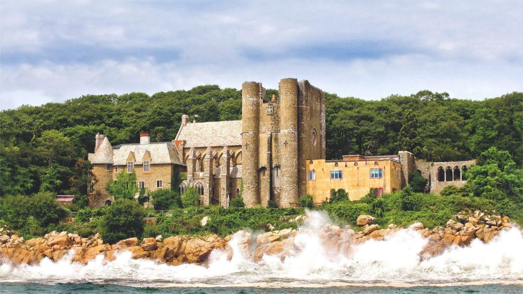 Stone castle ruins on a rocky coast, waves crashing below, surrounded by green trees and a cloudy sky.