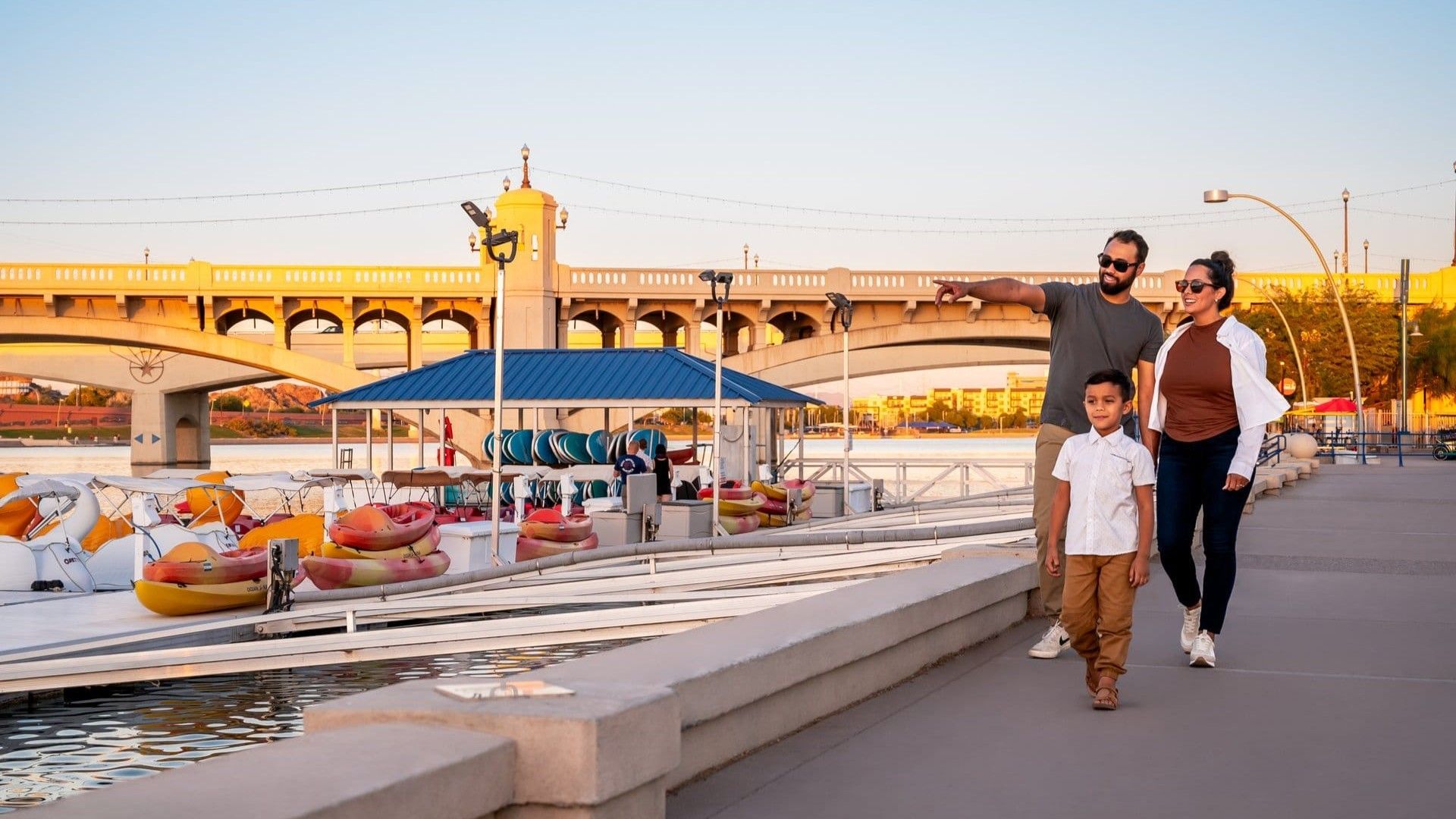 Family walking on a waterfront path, pointing toward the water, with a bridge in the background.