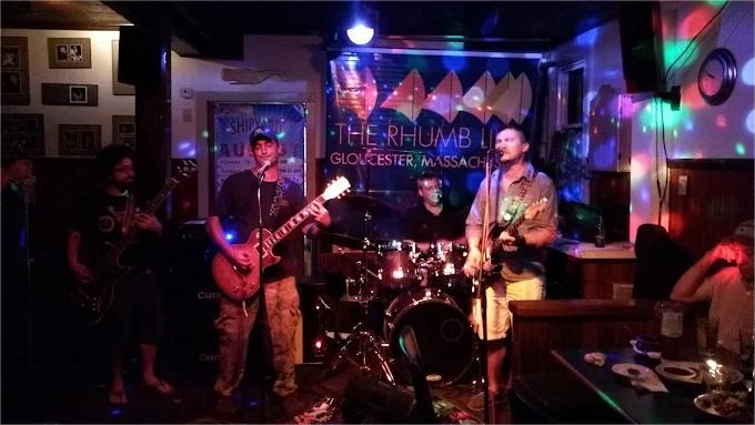 Band playing on stage at a bar: musicians with instruments, stage lights, and a backdrop banner.