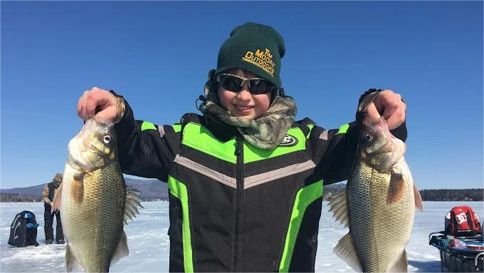 Boy ice fishing, holding up two fish on a frozen lake, wearing winter gear, sunny day.