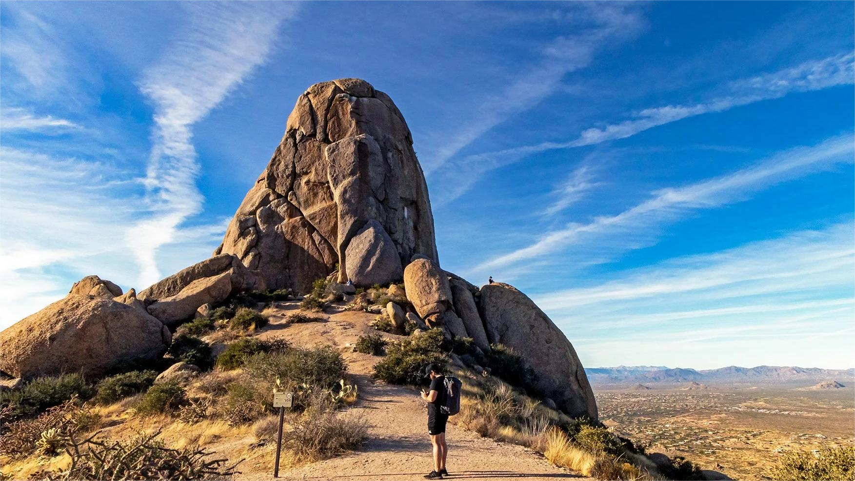 Man hiking on a trail towards a large rock formation under a blue sky, desert landscape.