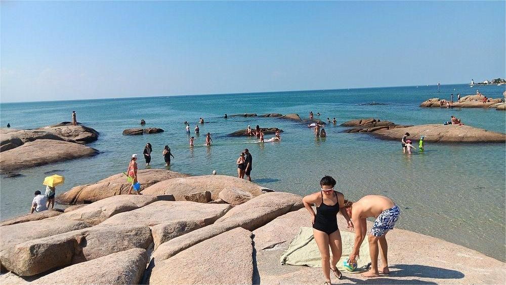 People swimming and sunbathing at a rocky beach with clear blue water under a clear sky.