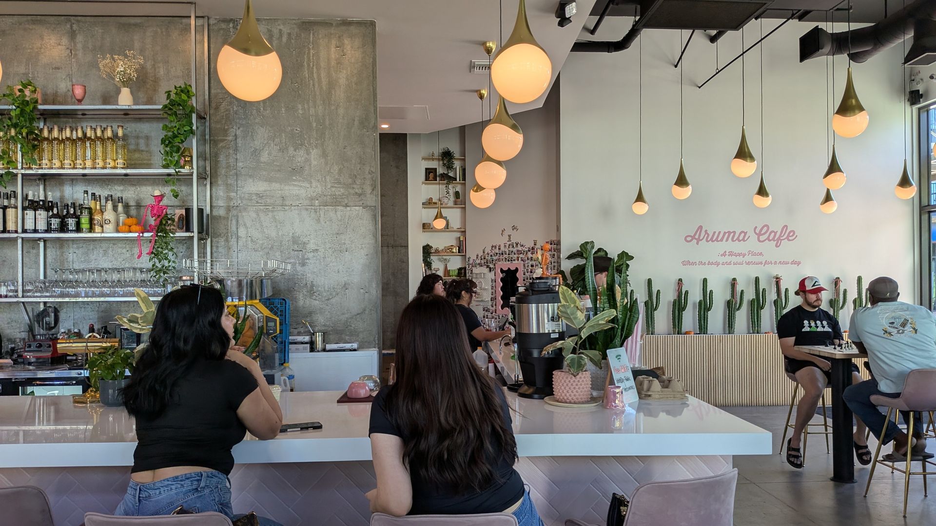 Interior of a cafe with people at the bar, overhead lights, a concrete wall, and cacti decor.