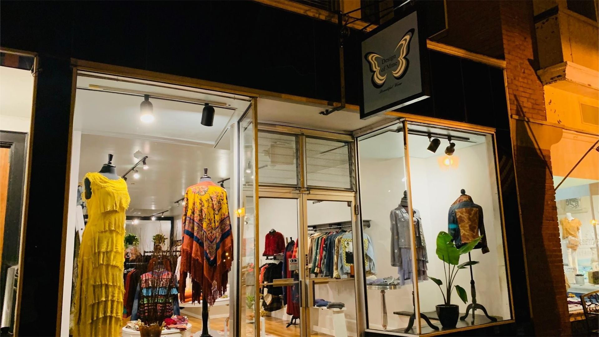 A boutique storefront at night, displaying clothing on mannequins and racks under warm lighting. Sign above shows a butterfly.