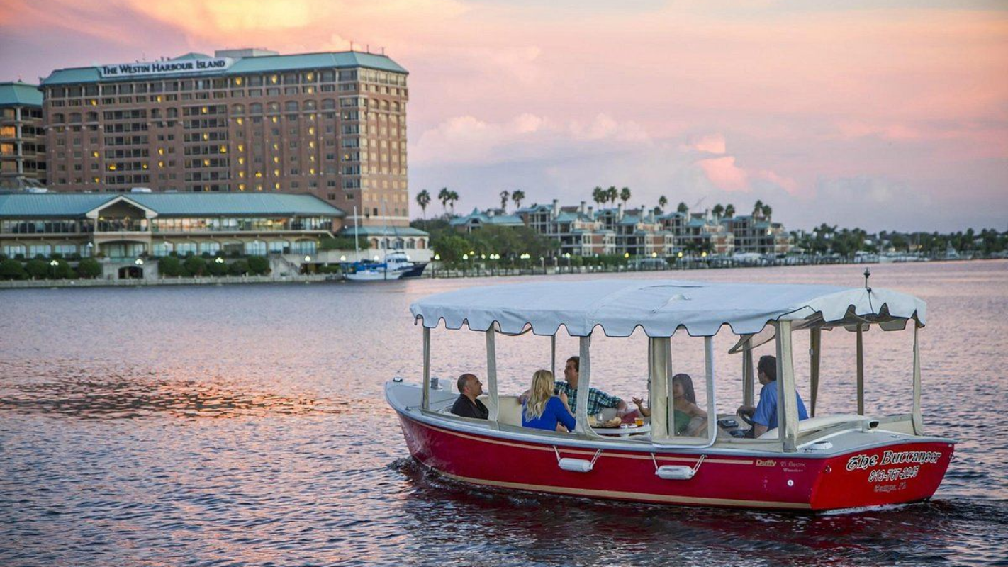 Red boat with passengers on water, buildings in background at sunset.