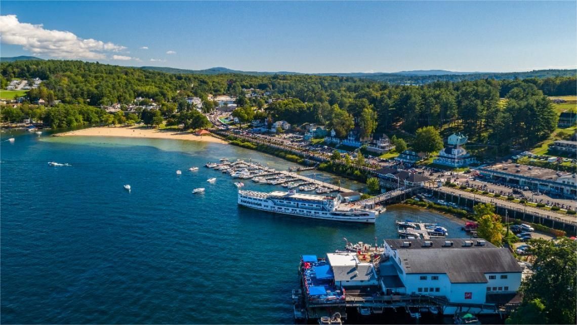 Aerial view of a coastal town with a marina, beach, and forested hills under a blue sky.