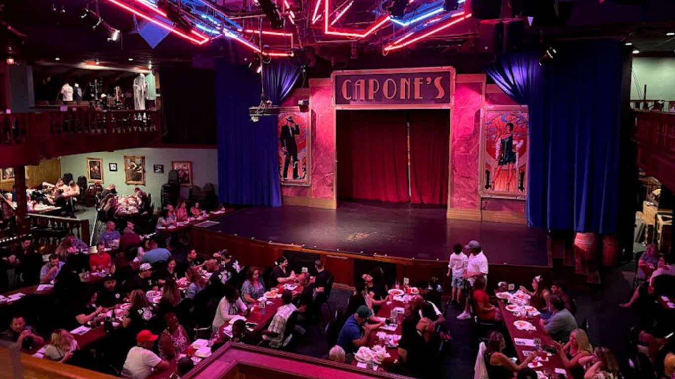 Interior of Capone's dinner theater; stage with curtains, audience at tables, red lighting.