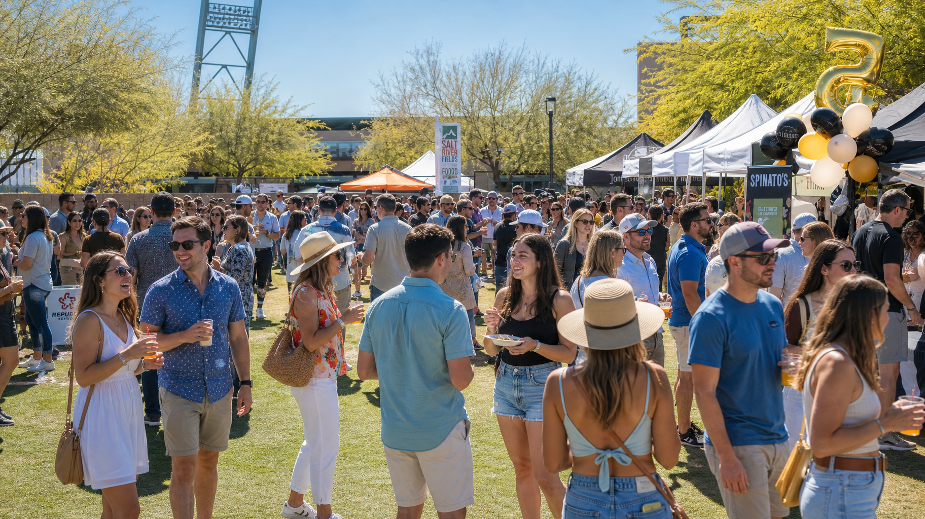 Crowded outdoor festival with people mingling near white tents and black-and-gold balloons on a sunny day
