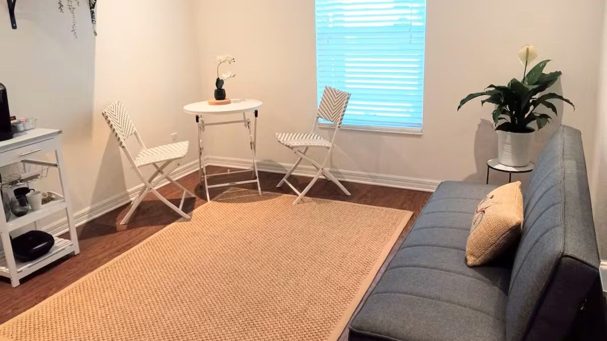 Small, furnished room with a gray sofa, table, chairs, rug, and potted plant. Beige walls, wooden floor, and a window.