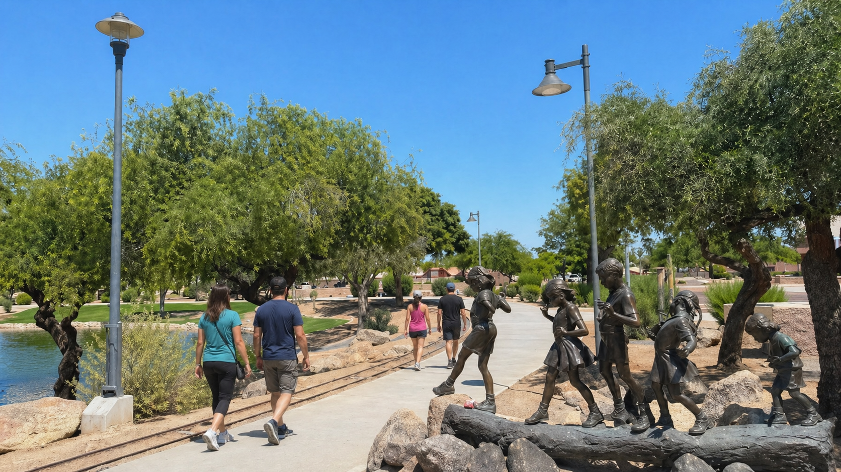 People walking along a sunny park path beside trees, lamps, and a rocky fountain sculpture.