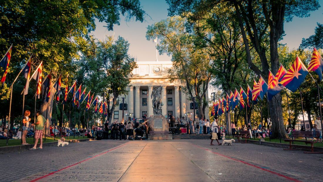 Flags line a brick pathway leading to a white building. People gather around a monument.