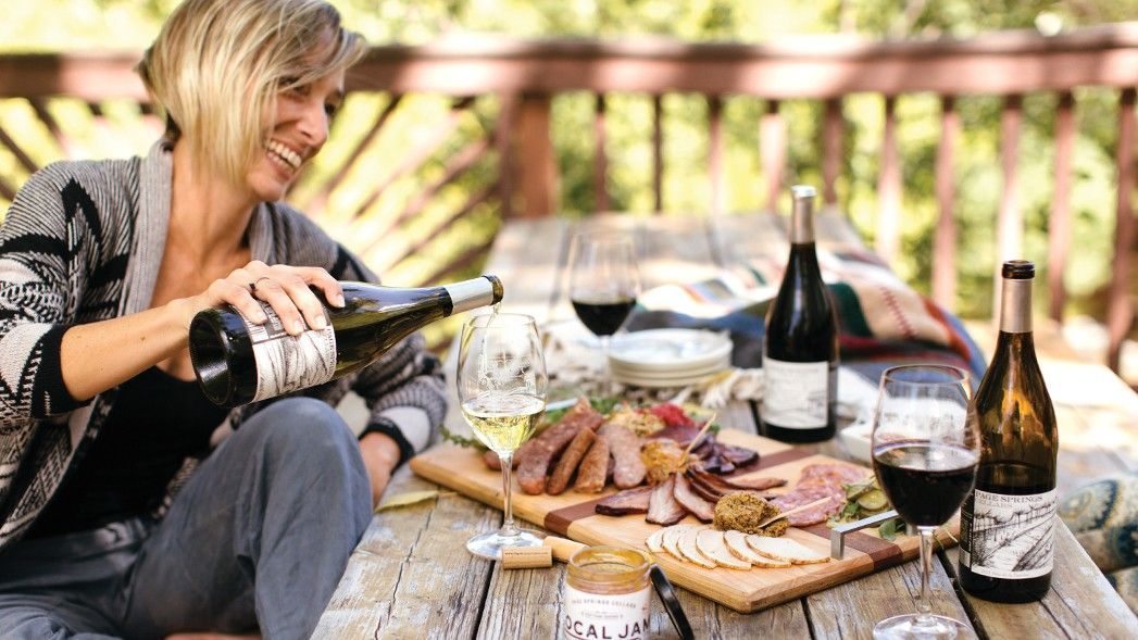 Woman pouring white wine at a picnic on a wooden deck. Charcuterie board, red wine, and bottles on table.
