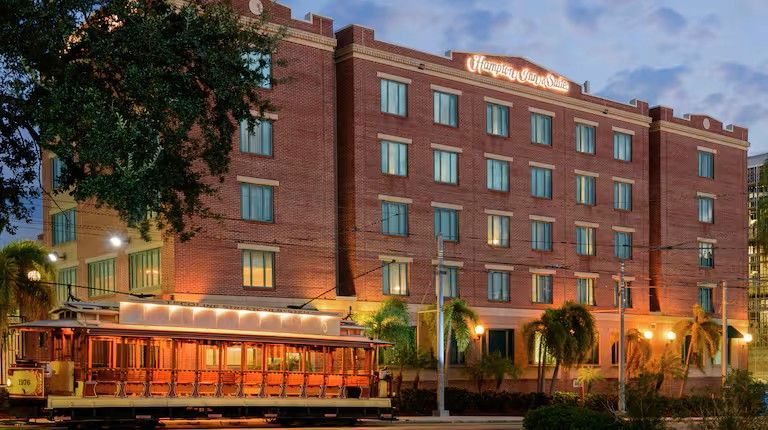 Brick hotel with Hampton Inn signage, trolley car, trees, and street lights at dusk.