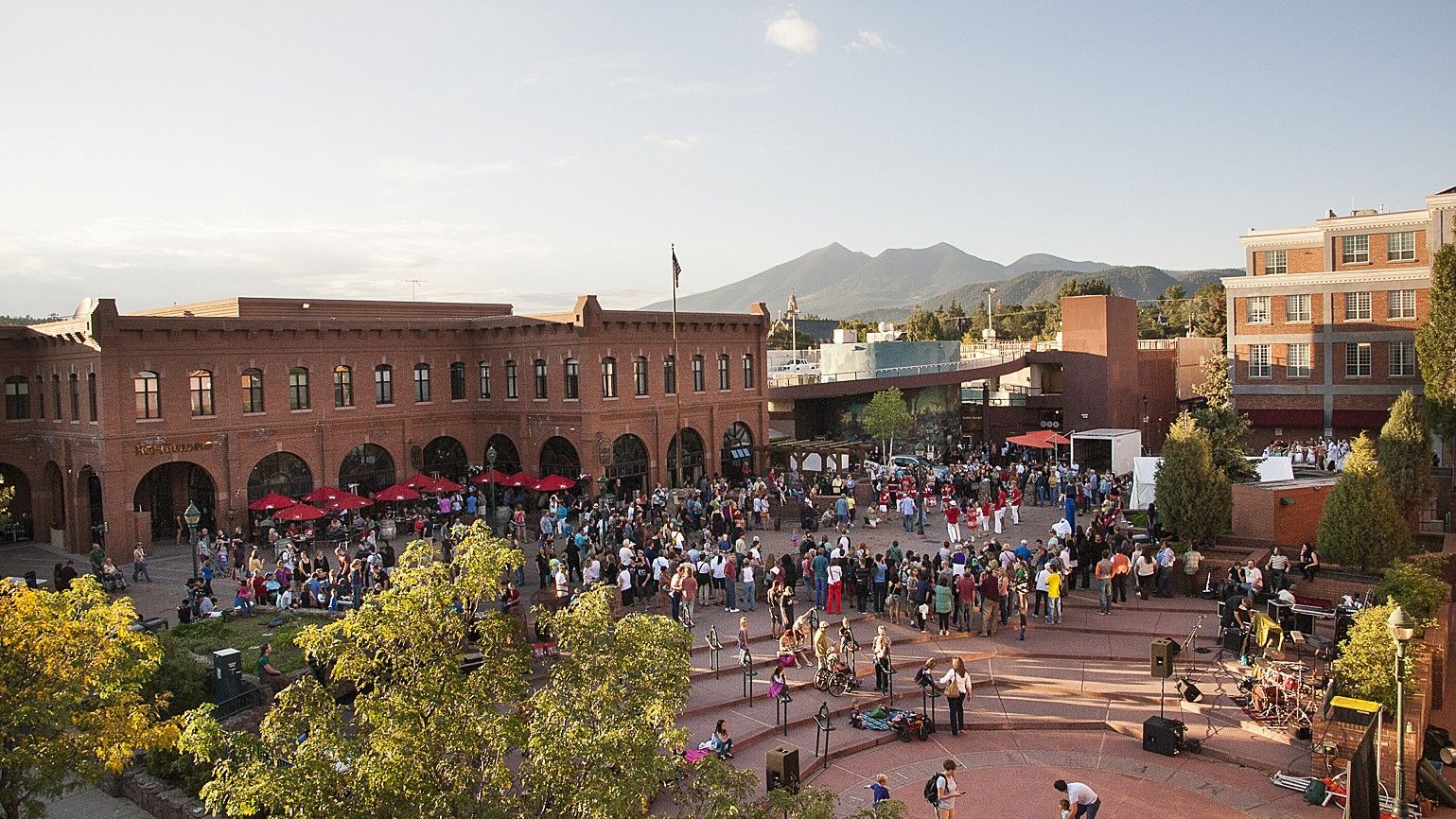 Large crowd in a plaza with brick buildings, trees, and mountains in the background.