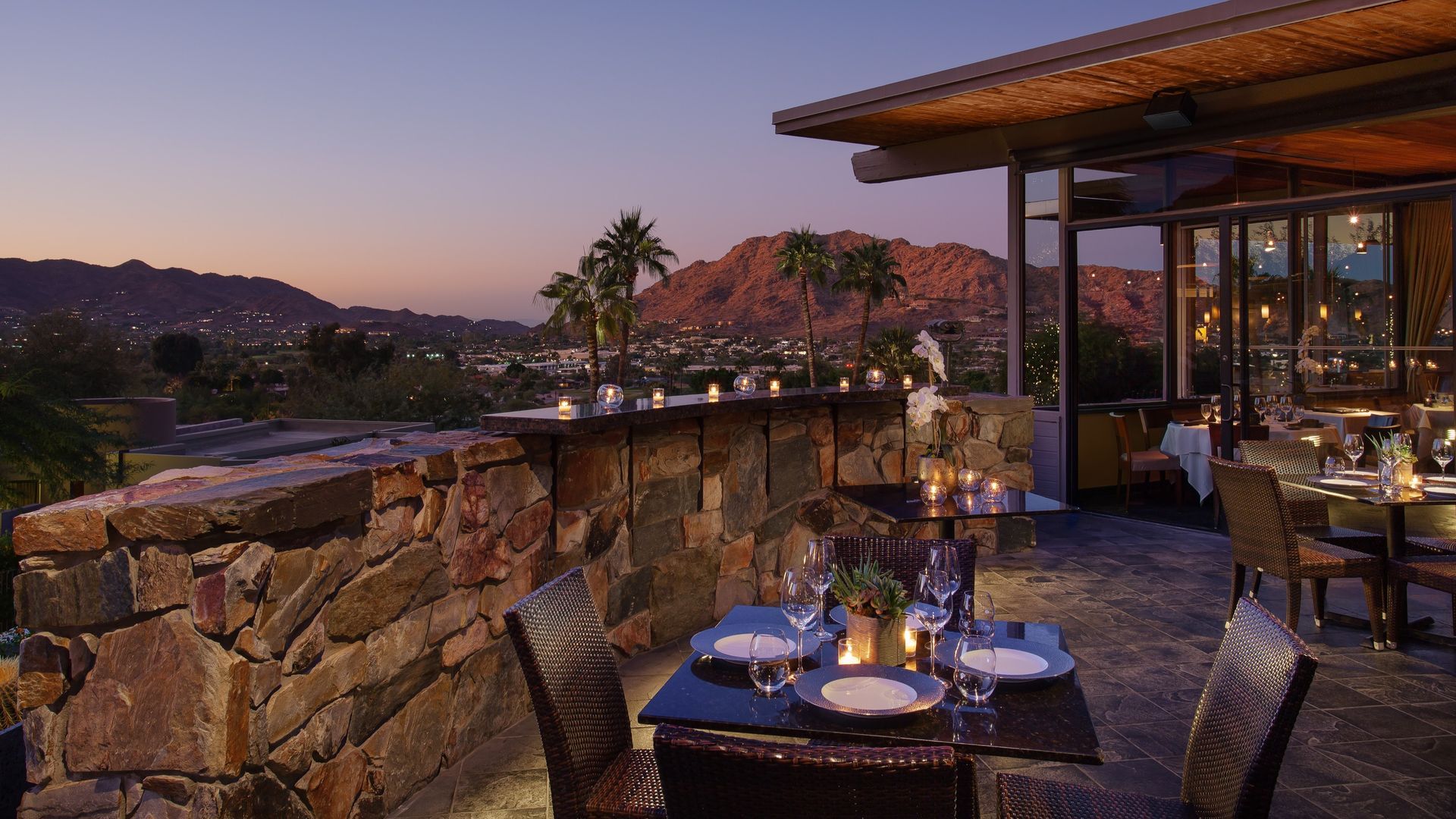 Outdoor restaurant patio at dusk overlooking a city and mountains. Table set for two with candle.