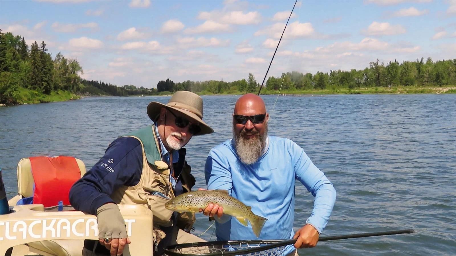 Two men on a boat, one holding a brown fish, river in background.
