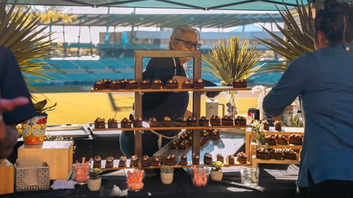 People arranging pastries on a tiered display at an outdoor event, stadium in the background.