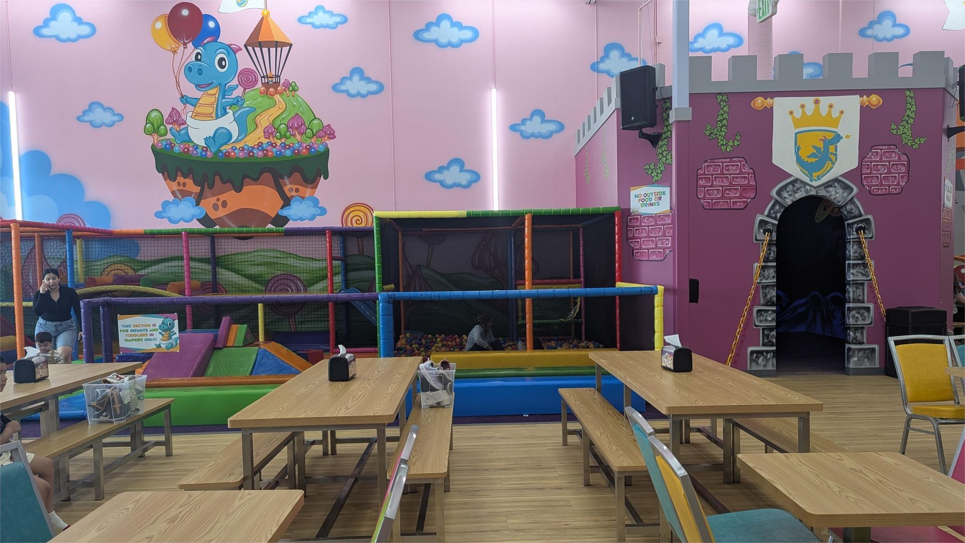 Indoor children's play area with tables, benches, and castle-themed structures. Pink walls, colorful mural.