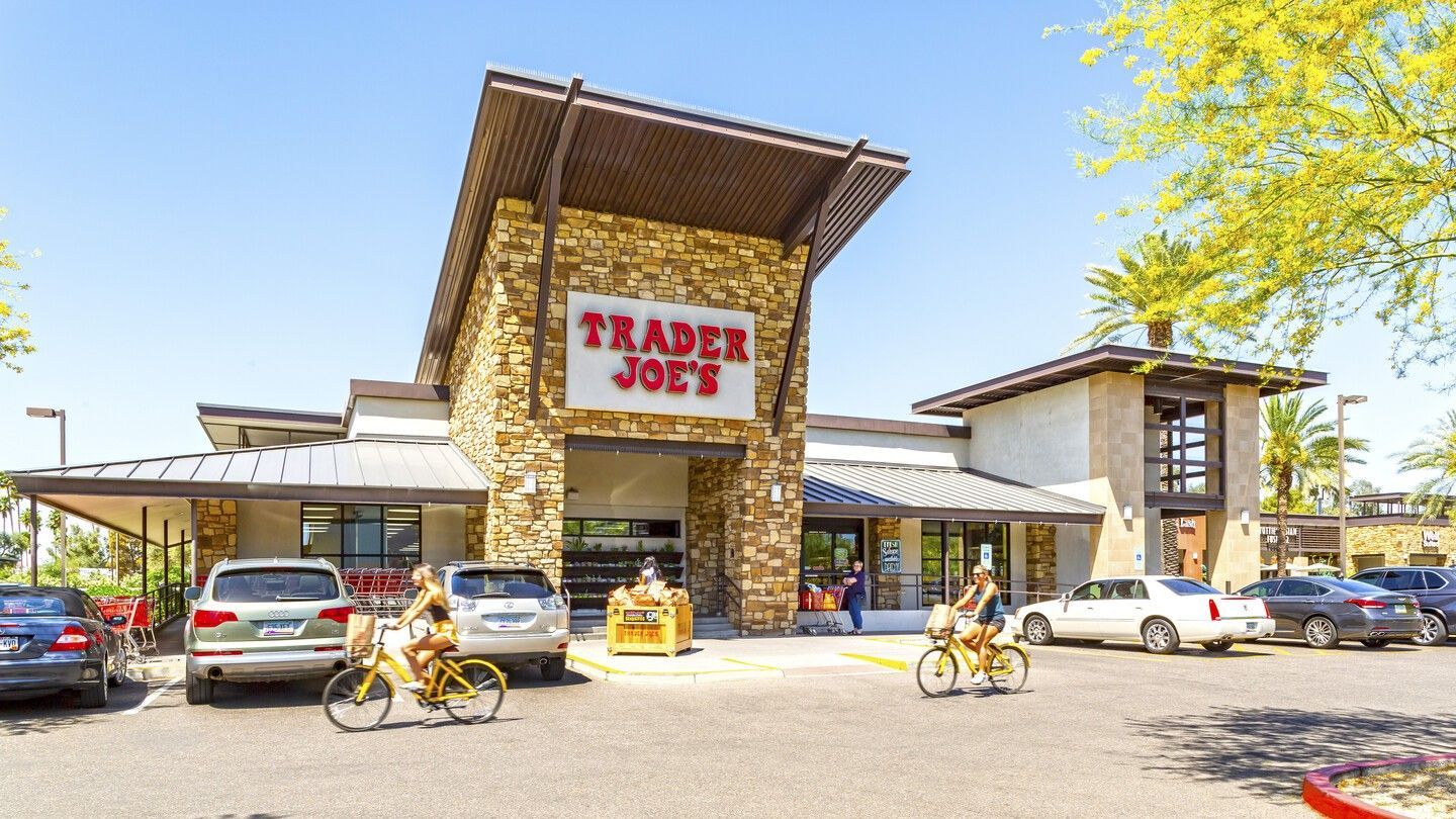 Trader Joe's store exterior with customers biking. Stone facade, parking lot, sunny day.