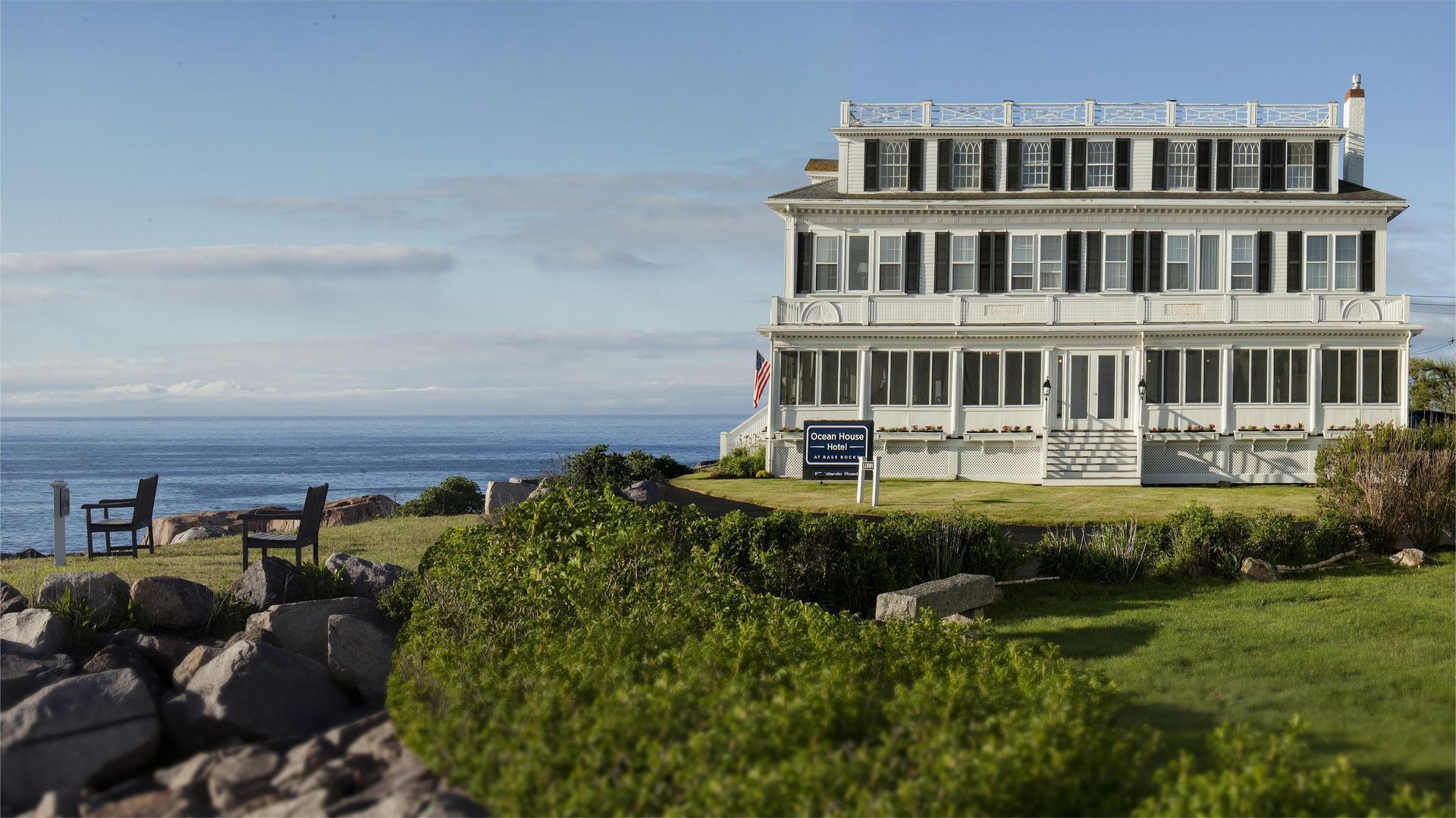 Large white multi-story house overlooking the ocean, with green grass and shrubbery in the foreground.