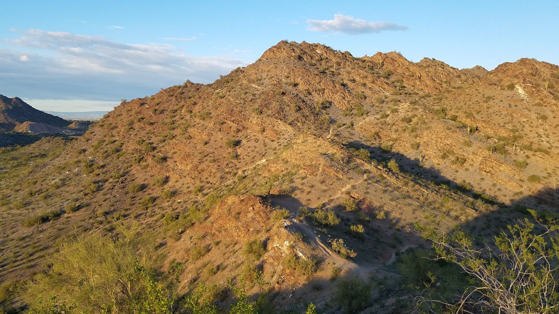 View of a rocky, sunlit mountain with a visible hiking trail under a blue sky.