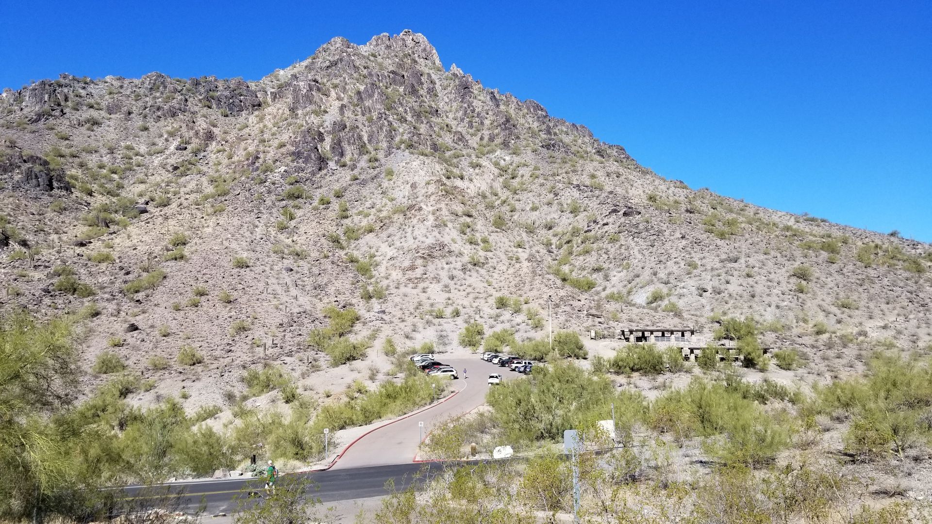 Mountain with a rocky peak and a winding road leading to a parking area. Blue sky.