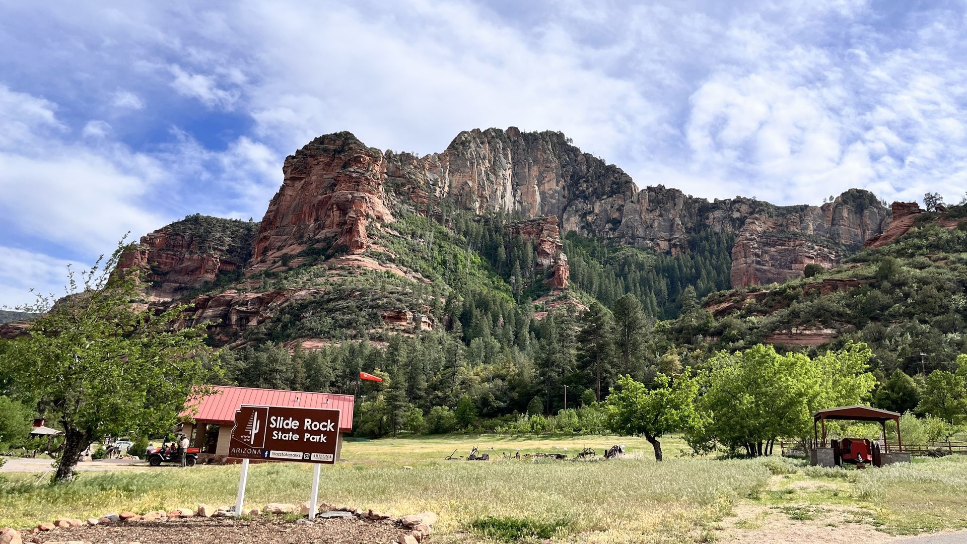 Mountain and field scene with a sign in the foreground and a house. Blue sky with clouds.