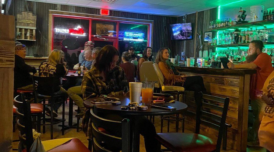 Bar interior with patrons seated at tables and bar. Neon lights illuminate the shelves with bottles.
