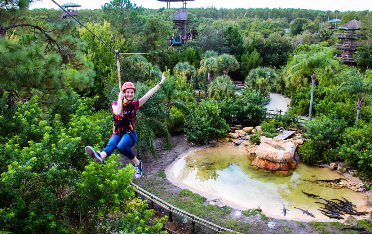 Woman ziplining through trees, arms raised, over a pond with a rock formation and dense foliage.