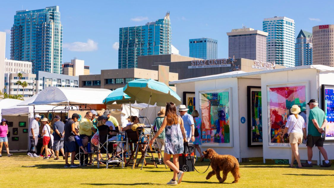 Art fair in a park, people viewing art displayed in booths, city skyline backdrop.