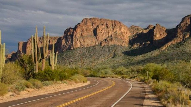 Desert road curves toward a rocky mountain under a cloudy sky, with cacti and scrub brush.