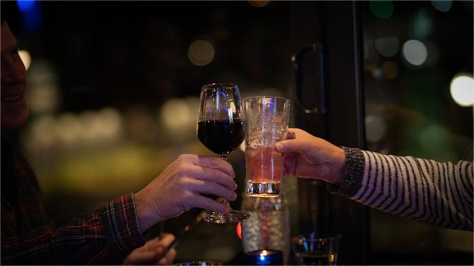 Two people toasting drinks at a bar, one holding red wine, the other a cocktail. Dark setting, blurred lights.