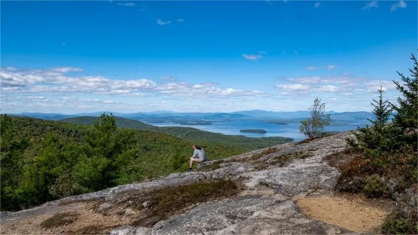 Person on rocky mountaintop overlooks a lake and distant hills under a bright blue sky.