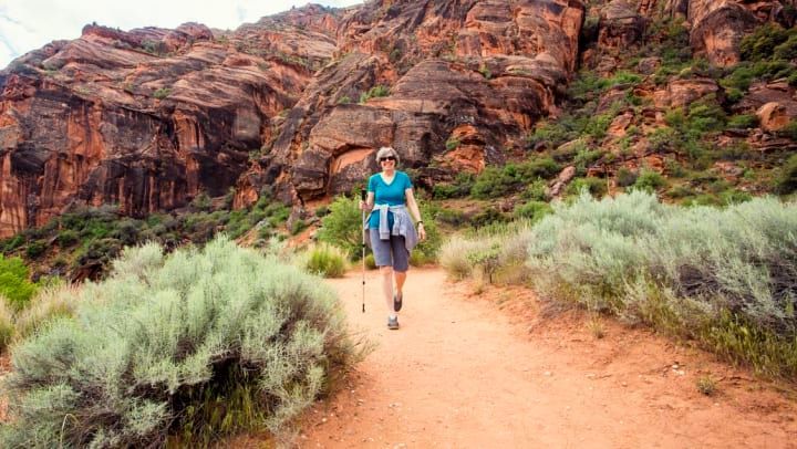 Woman hiking on a red dirt trail, surrounded by green shrubs, with red rock canyon in the background.