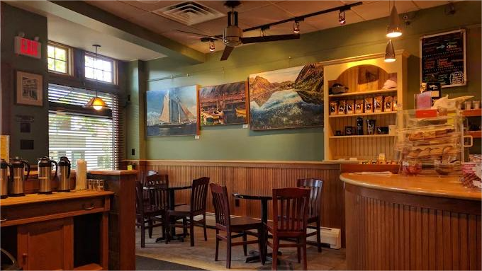 Interior of a coffee shop: wooden tables and chairs, artwork on wall, counter with pastries, coffee pots.