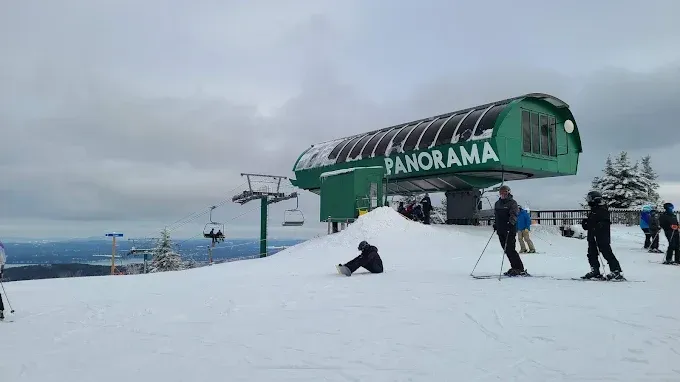 Panorama ski lift station on a snowy mountain, skiers and snowboarders nearby under a cloudy sky.