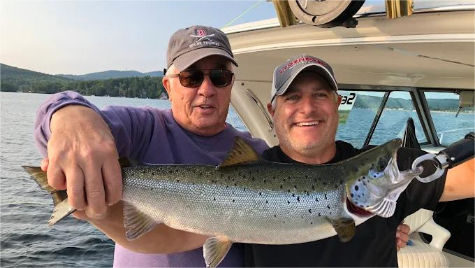 Two men on a boat hold up a large fish they caught, smiling. They are on a lake, with mountains in the background.